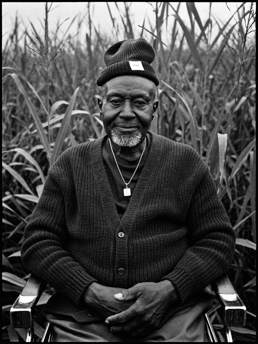 An elder in a black knit cardigan and beanie seated in a maize field